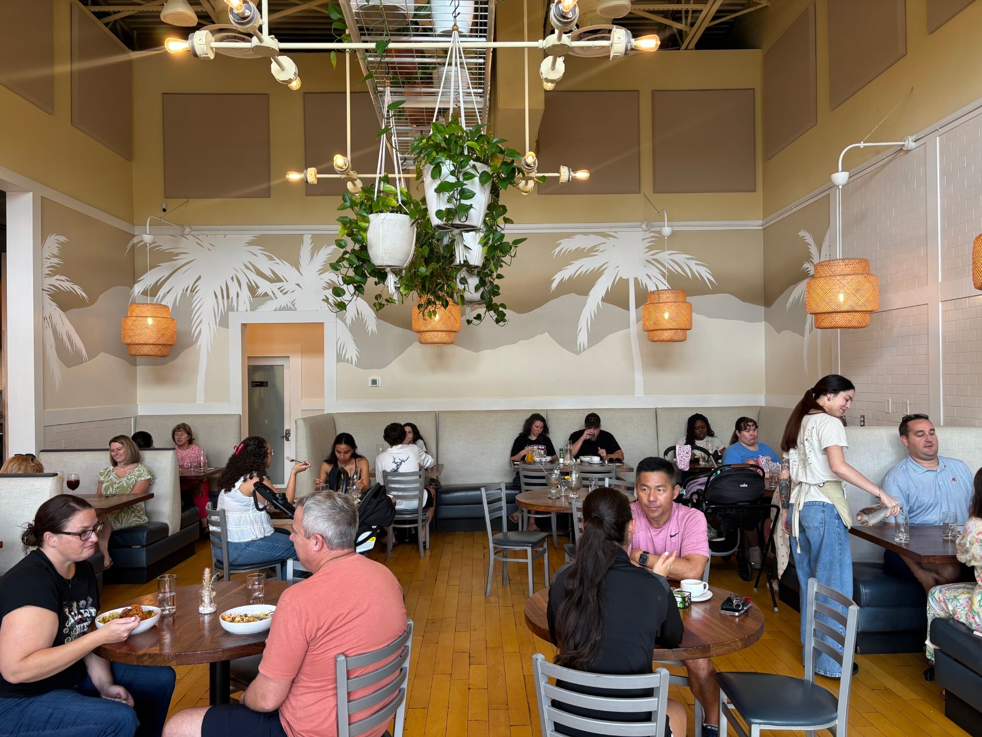 Guests dining at Cali in Winthrop beneath a palm-silhouette mural, hanging potted plants, and wicker pendant lights in the restaurant's current dining room