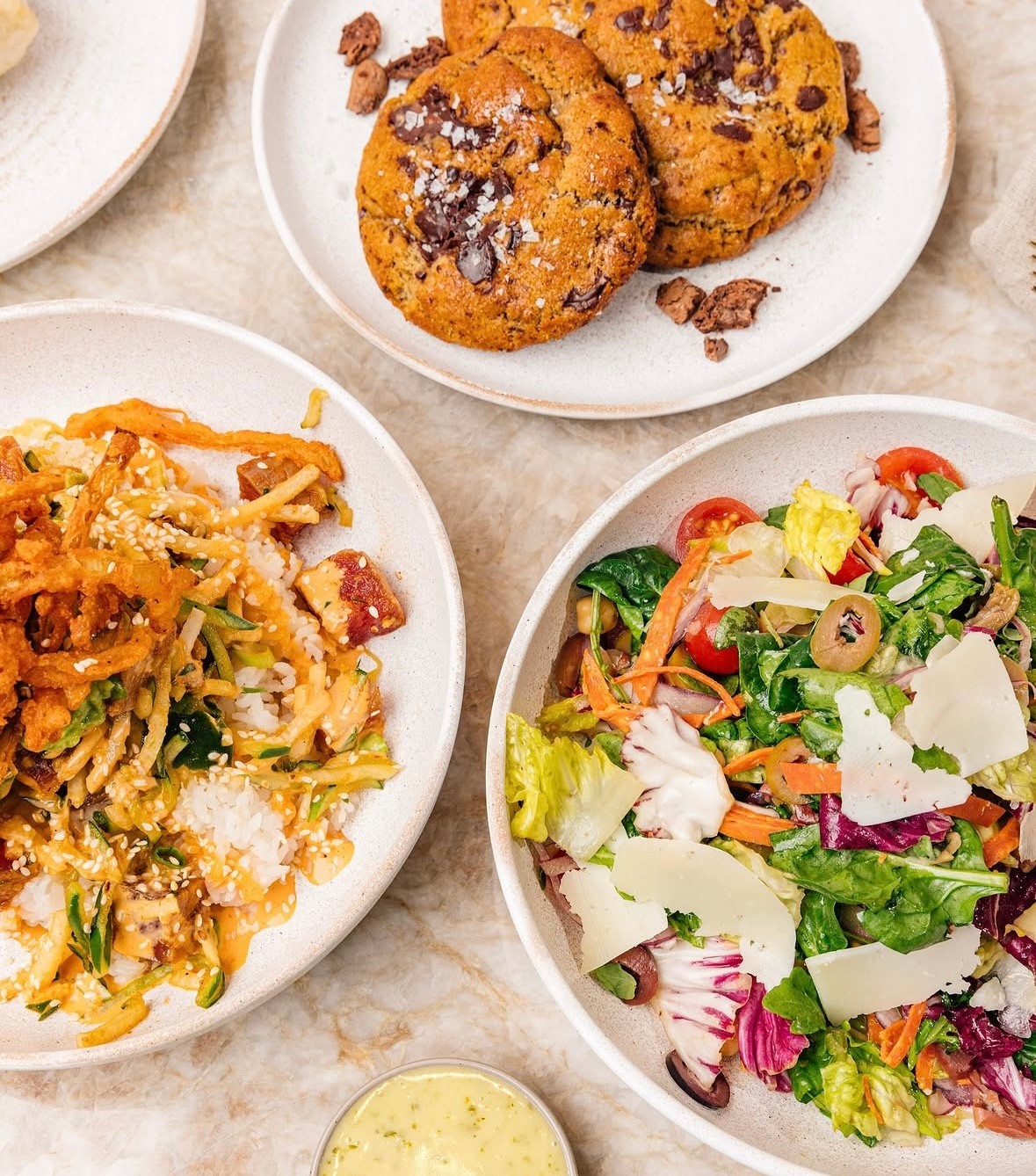 A flat-lay of Cali dishes — a mixed greens salad with tomatoes, olives, and shaved cheese, a rice bowl with crispy sesame toppings, and chocolate chunk cookies with sea salt
